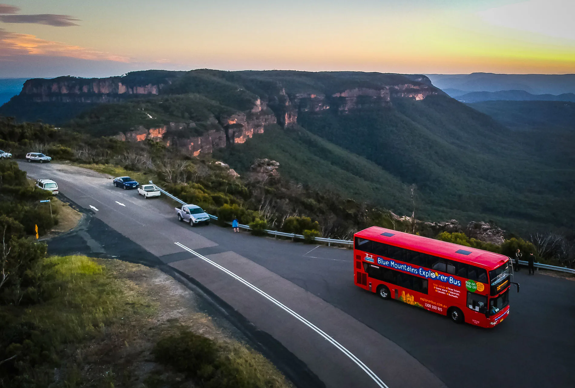 A red double-decker bus travels along a road overlooking the expansive, tree-covered Blue Mountains at sunset. Several cars are parked on the roadside, with panoramic views of the valleys and sandstone ridges in the background.