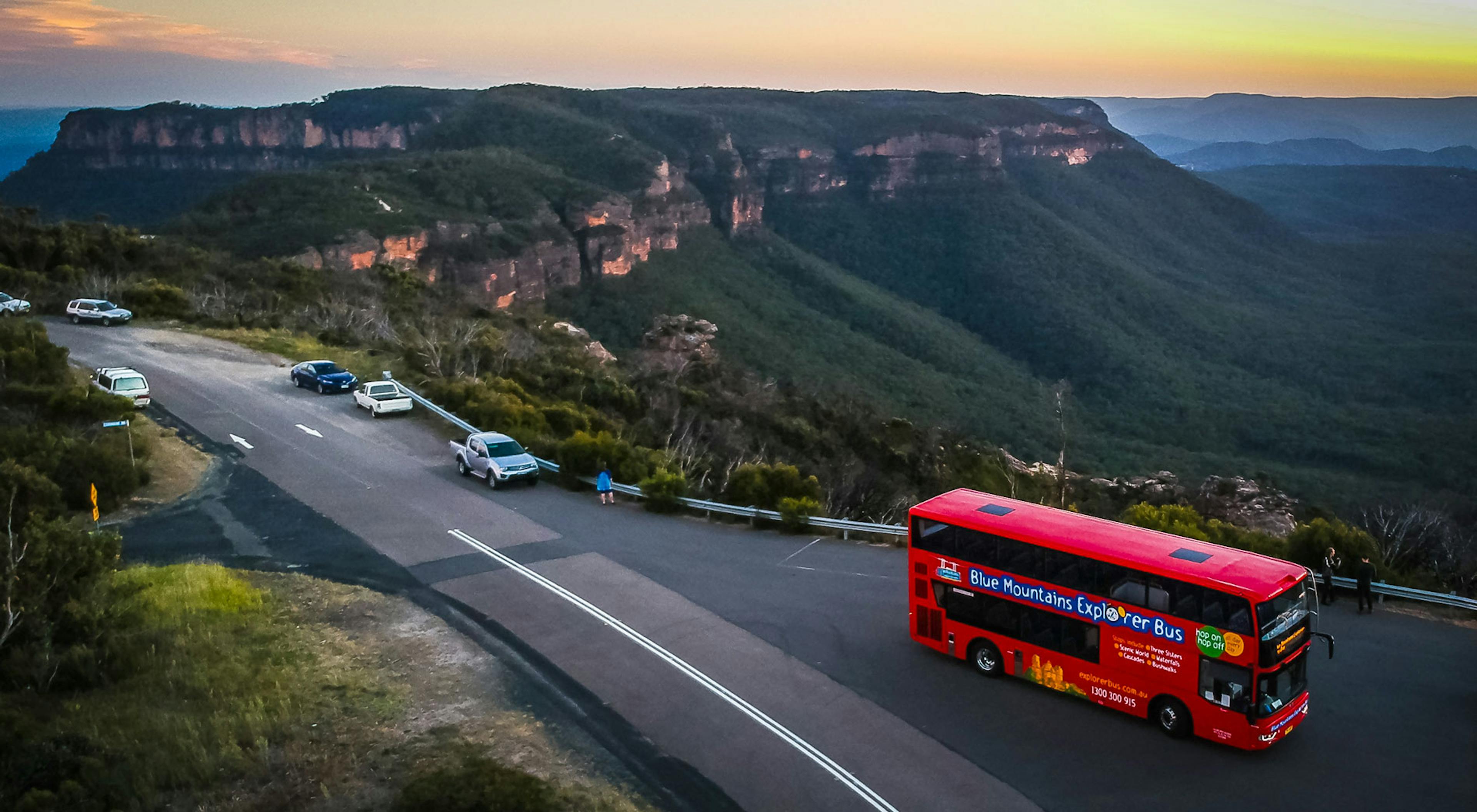 A red double-decker bus travels along a road overlooking the expansive, tree-covered Blue Mountains at sunset. Several cars are parked on the roadside, with panoramic views of the valleys and sandstone ridges in the background.