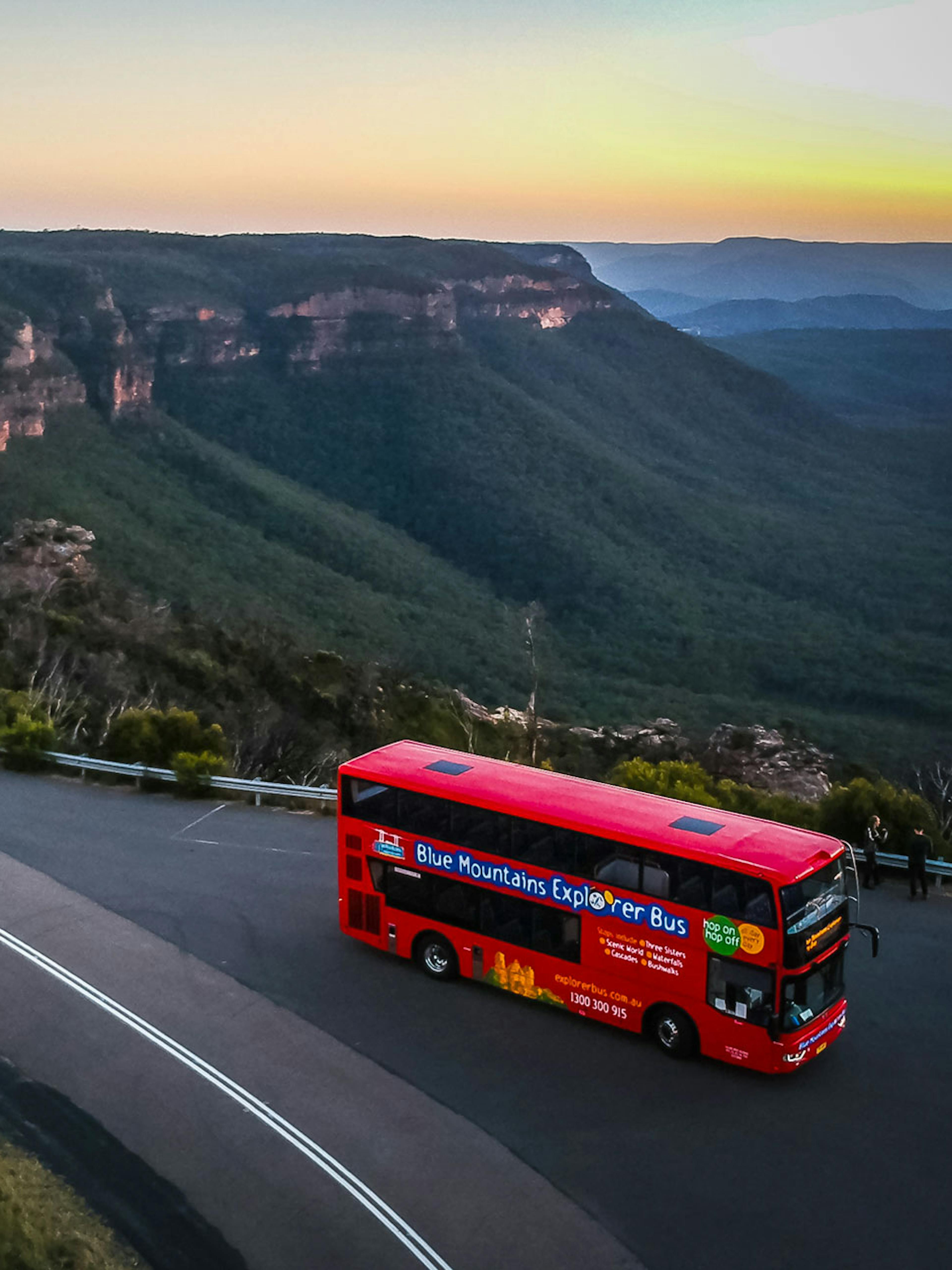 A red double-decker bus travels along a road overlooking the expansive, tree-covered Blue Mountains at sunset. Several cars are parked on the roadside, with panoramic views of the valleys and sandstone ridges in the background.