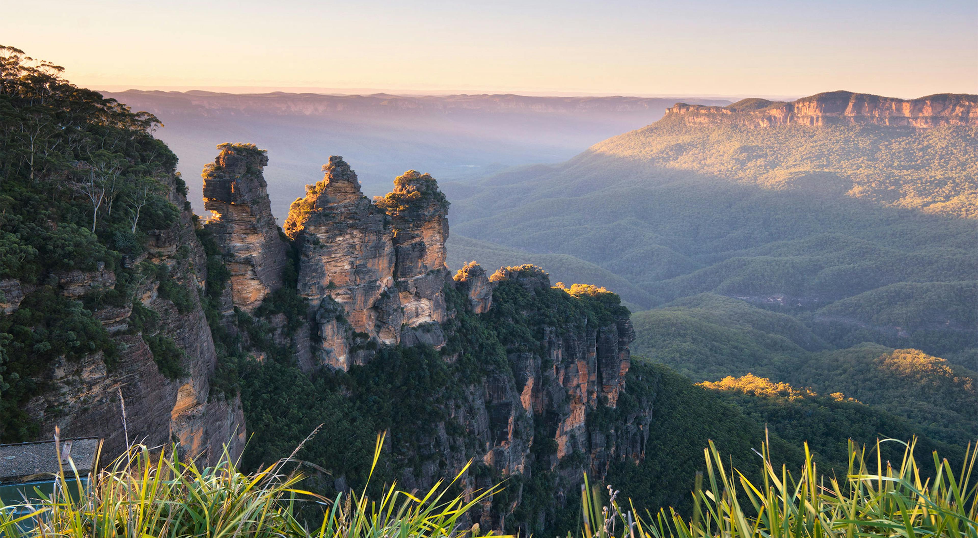 View of the Three Sisters rock formation at sunrise in the Blue Mountains, Australia. The peaks are surrounded by lush green valleys and distant mountains under a clear blue sky. Bright sunlight casts soft shadows across the landscape.