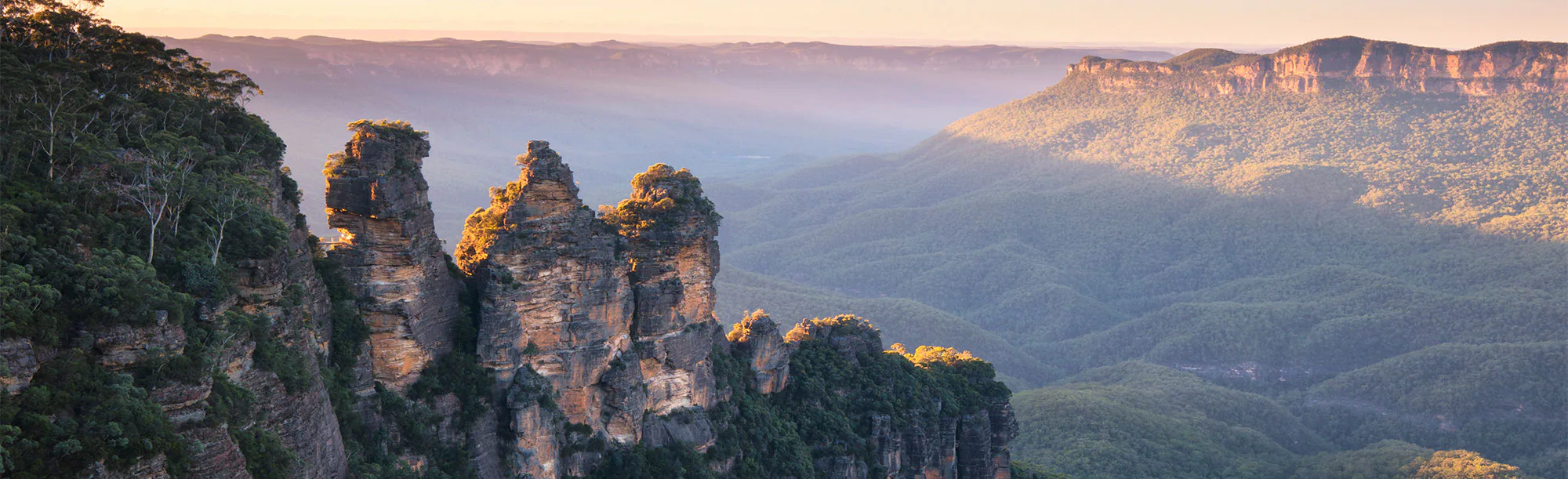 Scenic view of the Three Sisters rock formation at sunrise in the Blue Mountains, Australia. The peaks are surrounded by lush green valleys and distant mountains under a clear blue sky. Bright sunlight casts soft shadows across the landscape.