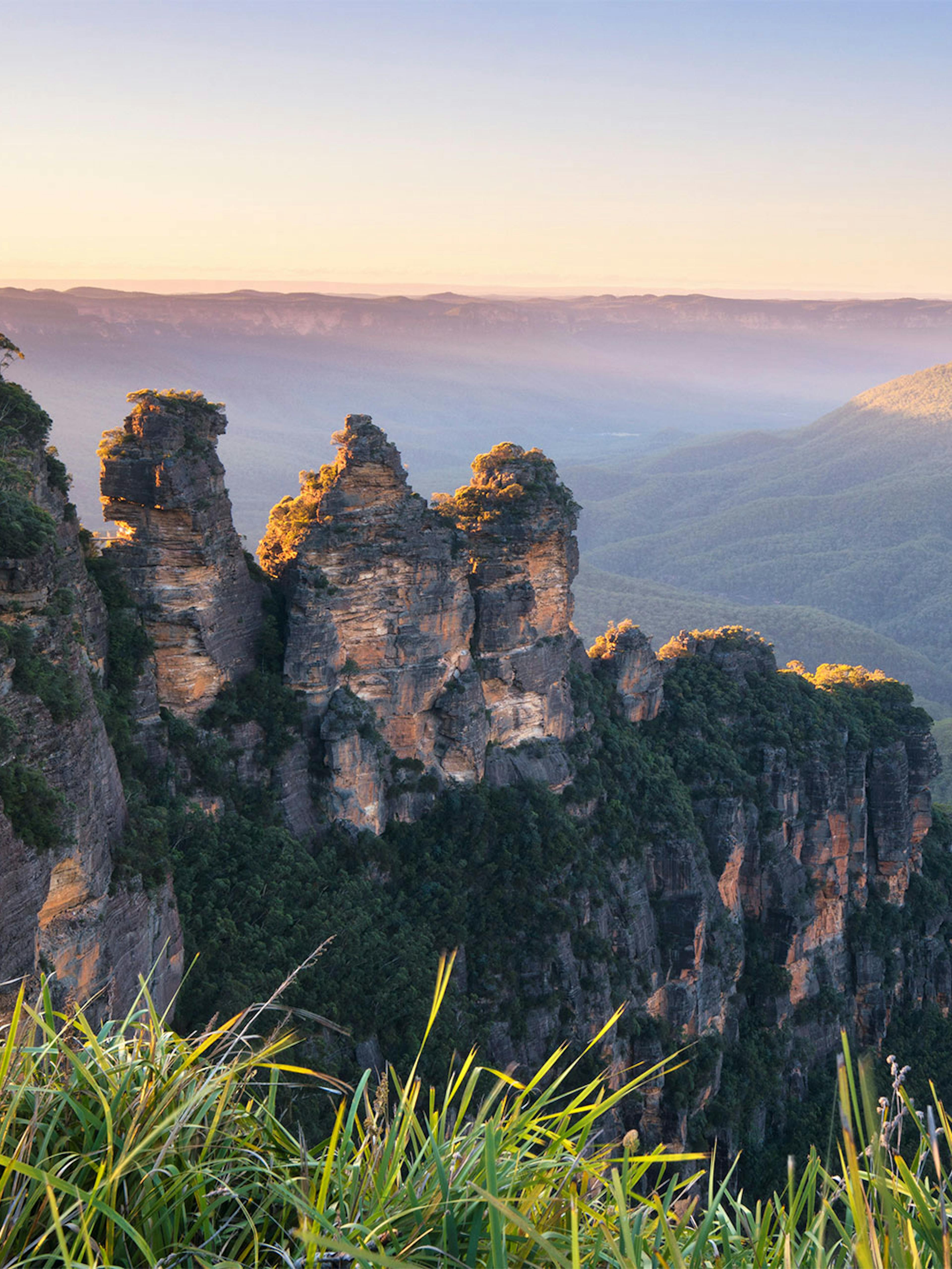 View of the Three Sisters rock formation at sunrise in the Blue Mountains, Australia. The peaks are surrounded by lush green valleys and distant mountains under a clear blue sky. Bright sunlight casts soft shadows across the landscape.