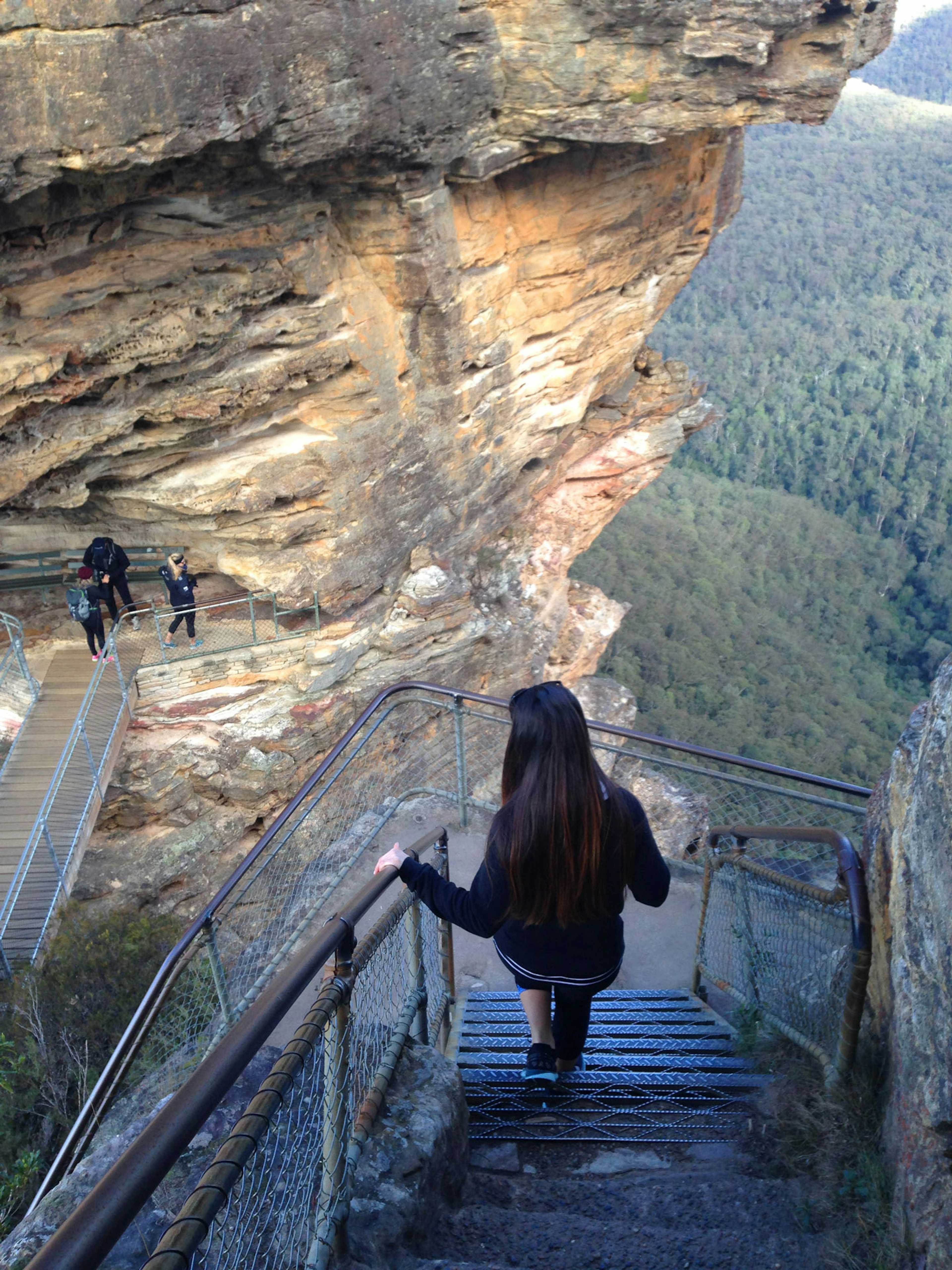 A woman descends a steep, narrow metal staircase carved into a rocky cliff. Two people ascend a bridge on the left. Dense forest covers a valley in the background under clear skies.