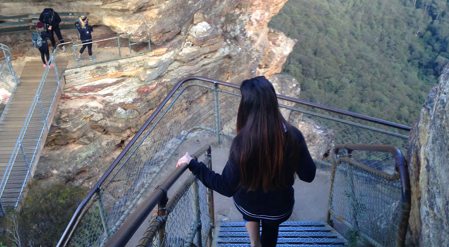 A woman descends a steep, narrow metal staircase carved into a rocky cliff. Two people ascend a bridge on the left. Dense forest covers a valley in the background under clear skies.