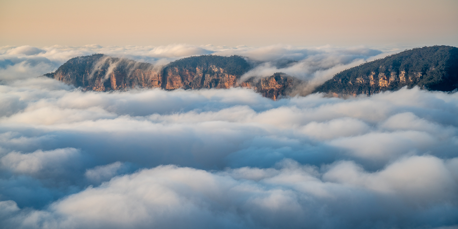 A scenic view of mountains partially covered by low-lying clouds or fog. The rugged peaks of the mountains emerge above the fluffy white clouds, with a soft, hazy sky in the background.