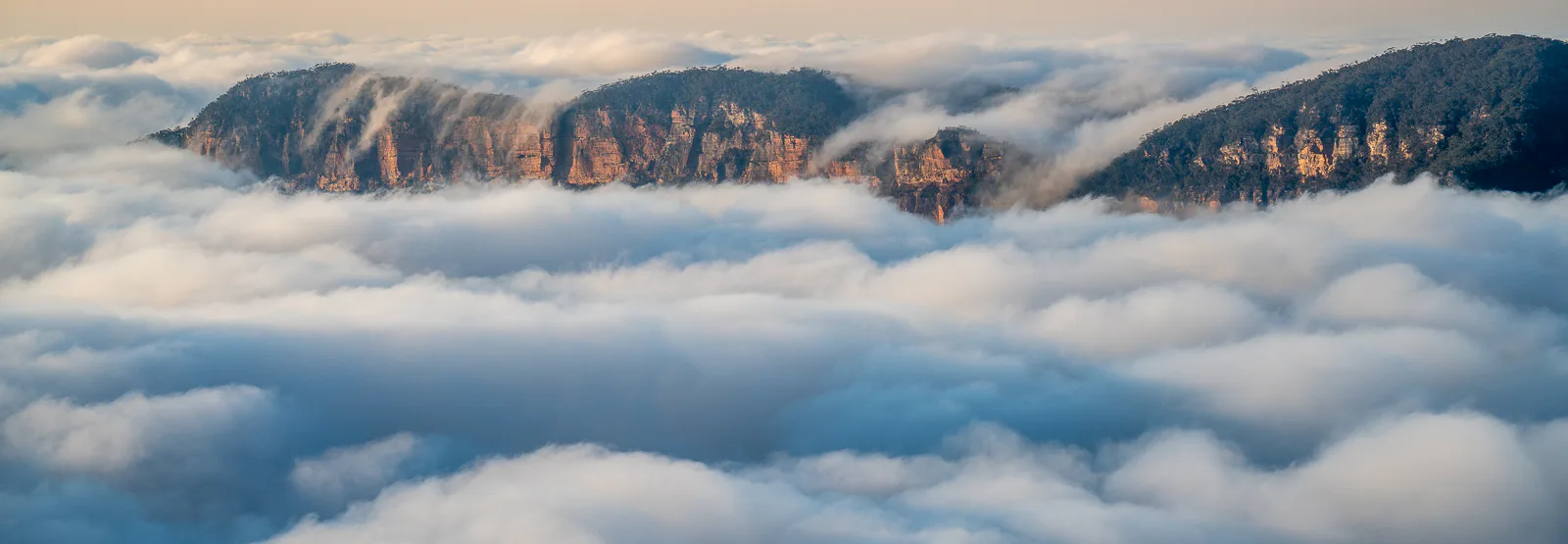 A scenic view of mountains partially covered by low-lying clouds or fog. The rugged peaks of the mountains emerge above the fluffy white clouds, with a soft, hazy sky in the background.