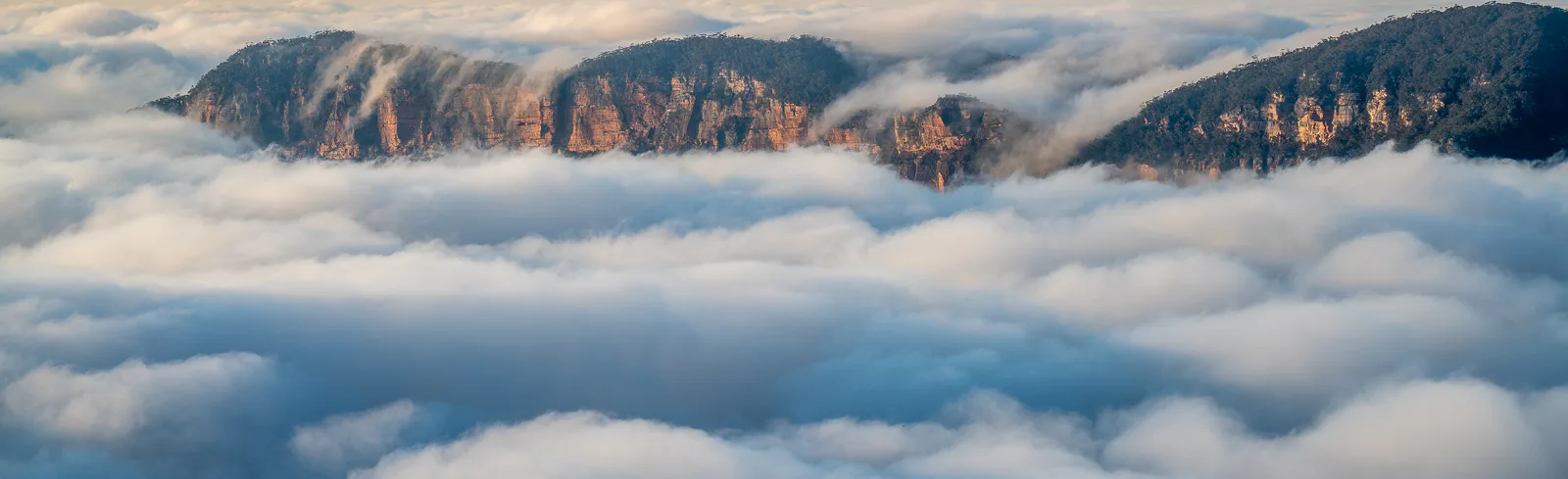 A scenic view of mountains partially covered by low-lying clouds or fog. The rugged peaks of the mountains emerge above the fluffy white clouds, with a soft, hazy sky in the background.