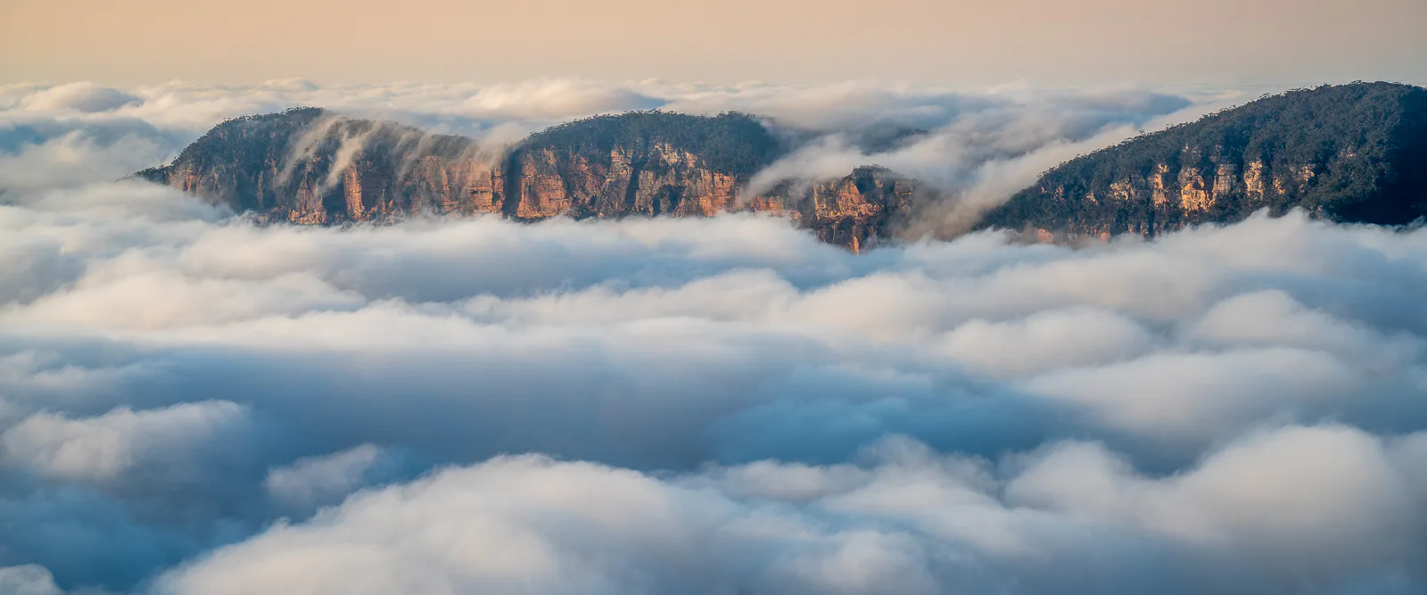 A scenic view of mountains partially covered by low-lying clouds or fog. The rugged peaks of the mountains emerge above the fluffy white clouds, with a soft, hazy sky in the background.
