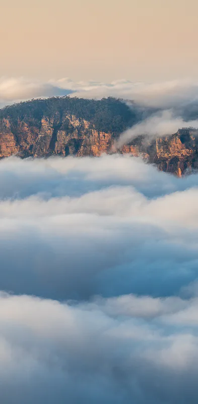 A scenic view of mountains partially covered by low-lying clouds or fog. The rugged peaks of the mountains emerge above the fluffy white clouds, with a soft, hazy sky in the background.