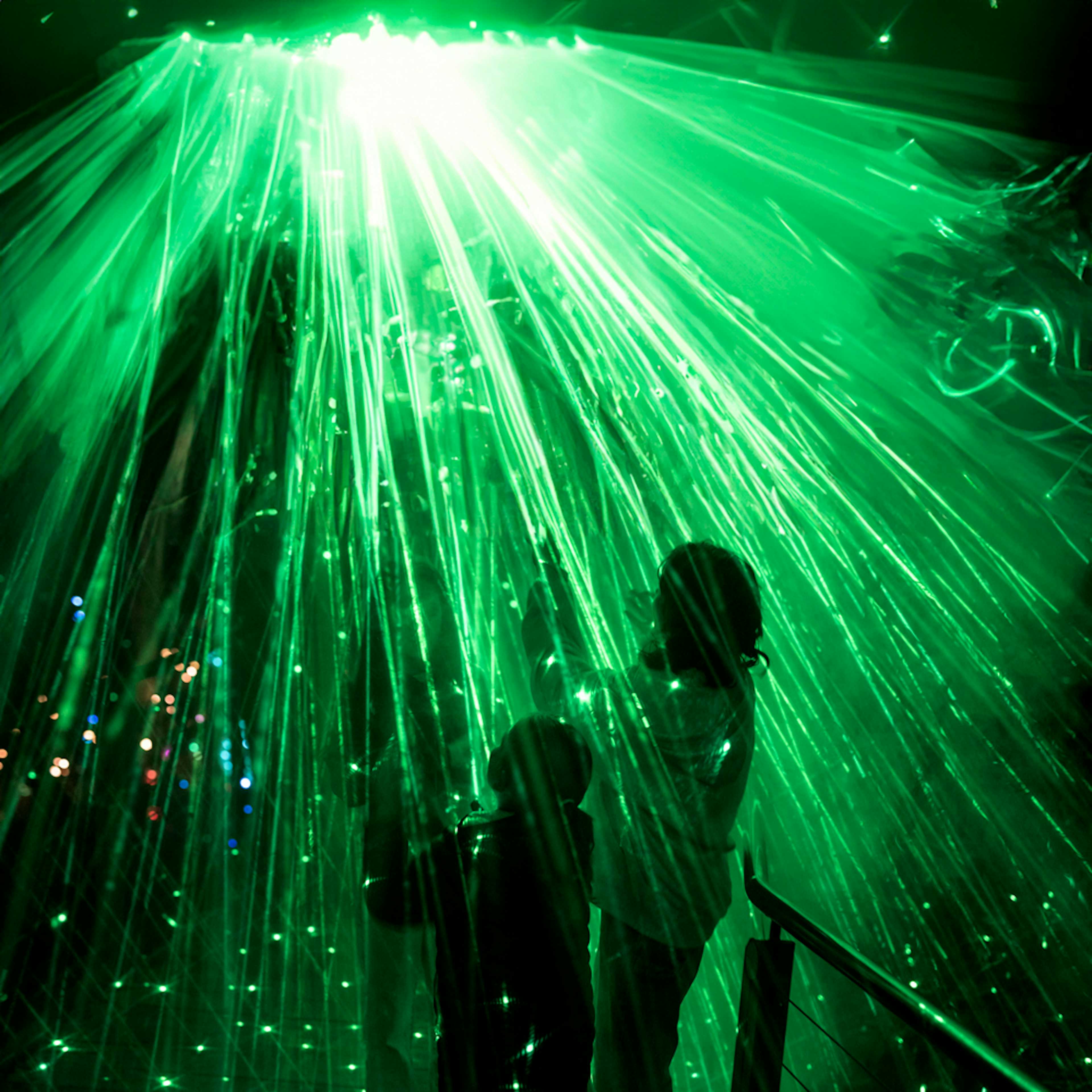 Silhouetted figures reach toward cascading green laser beams in a dark setting, creating a magical, otherworldly atmosphere.
