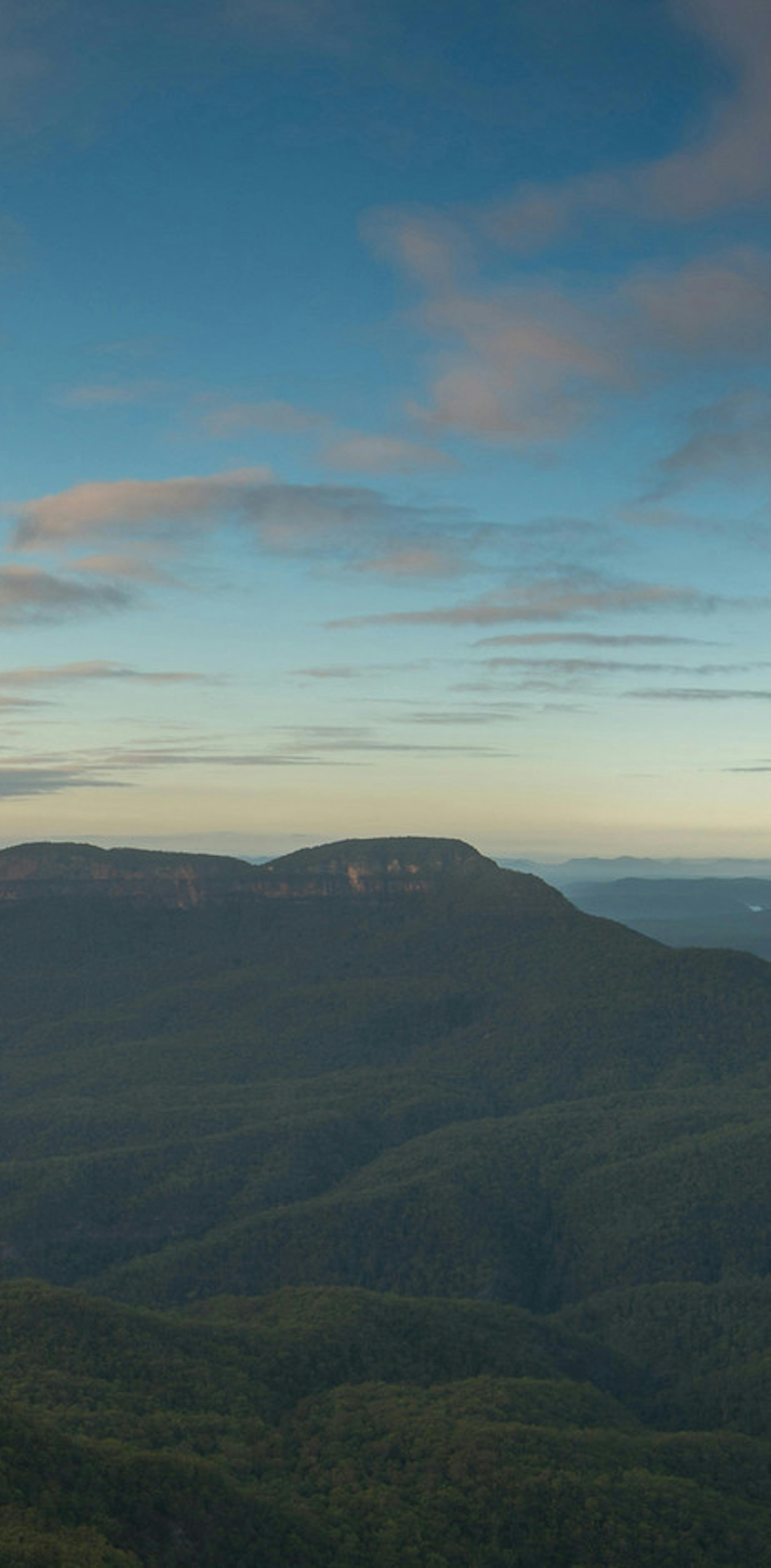 Panoramic view of a vast landscape with lush green valleys and rugged cliffs under a partly cloudy sky. The Three Sisters rock formation is prominently visible on the left, with sunlight illuminating the scene.