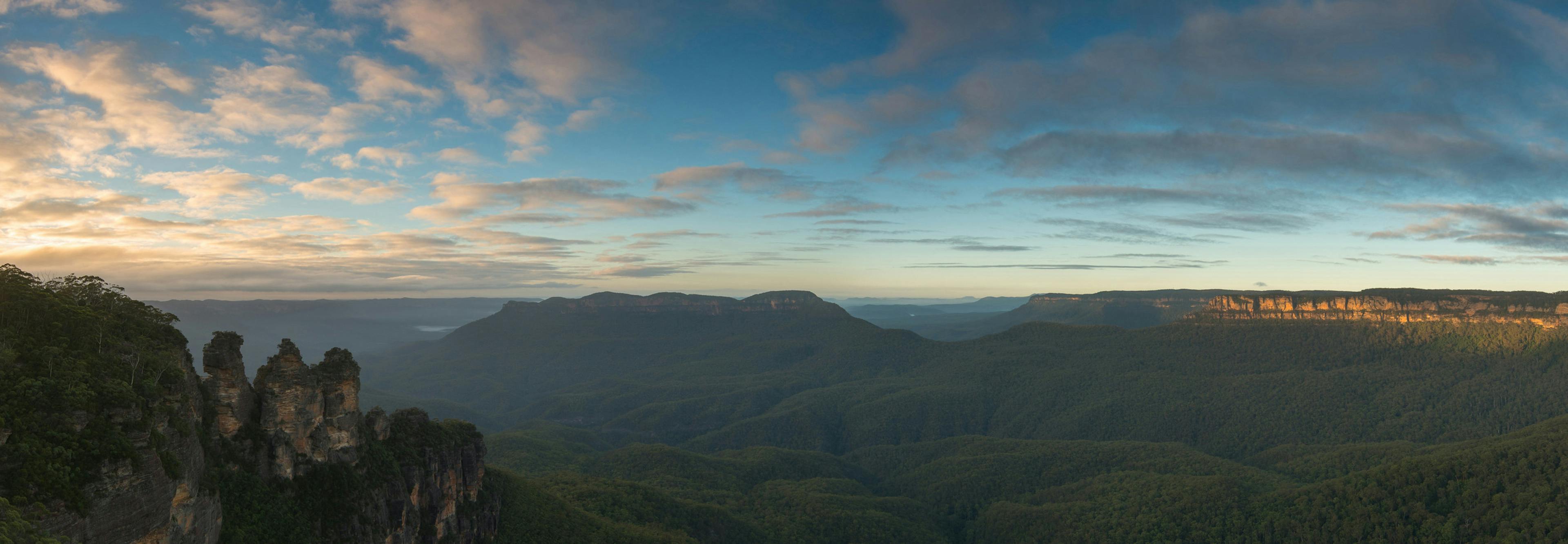 Panoramic view of a vast landscape with lush green valleys and rugged cliffs under a partly cloudy sky. The Three Sisters rock formation is prominently visible on the left, with sunlight illuminating the scene.