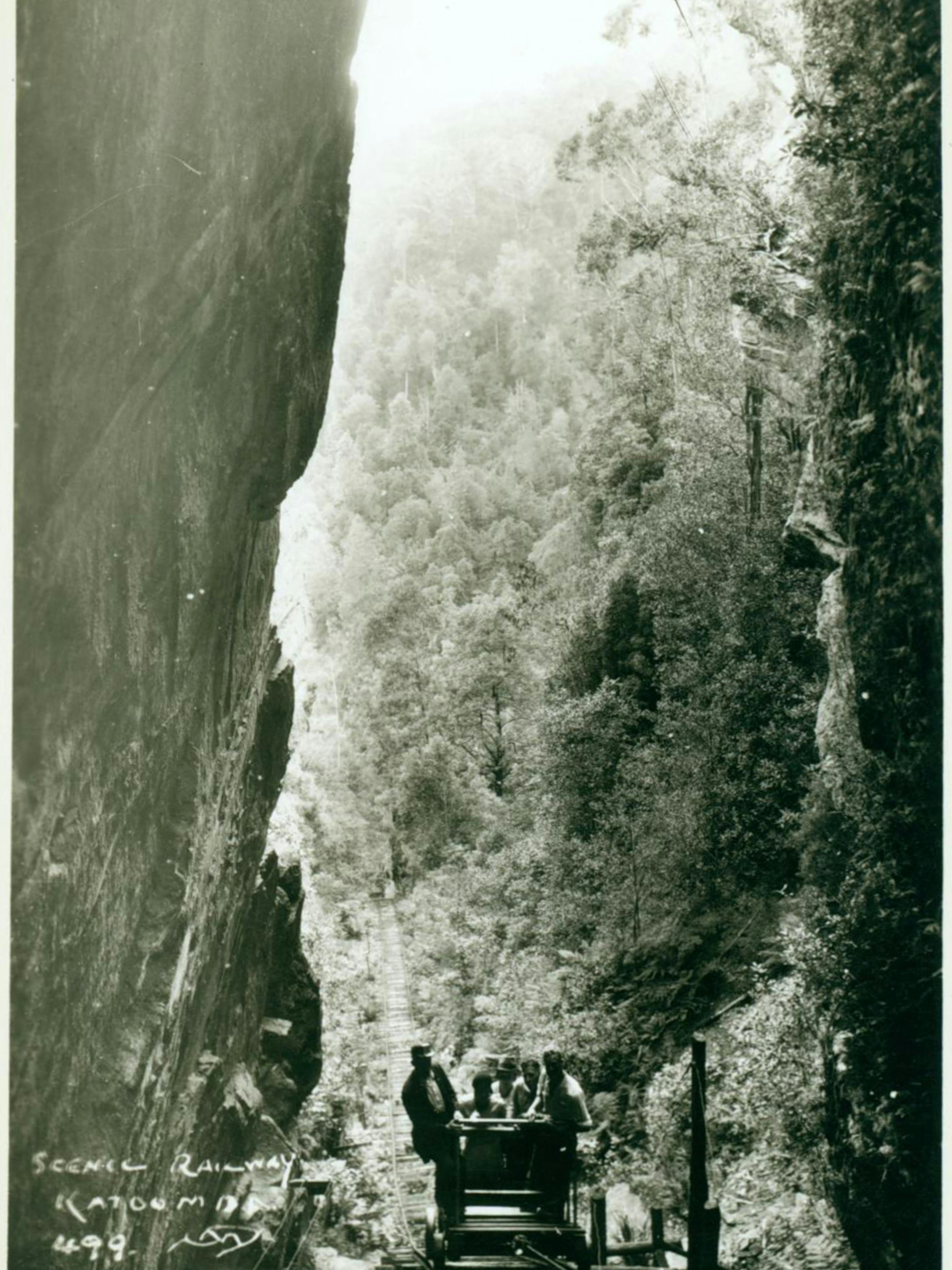 A vintage photograph shows a small group of people riding a scenic railway car through a narrow gorge surrounded by dense forest. The handwritten text Scenic Railway Katoomba 498 is visible in the lower left corner.