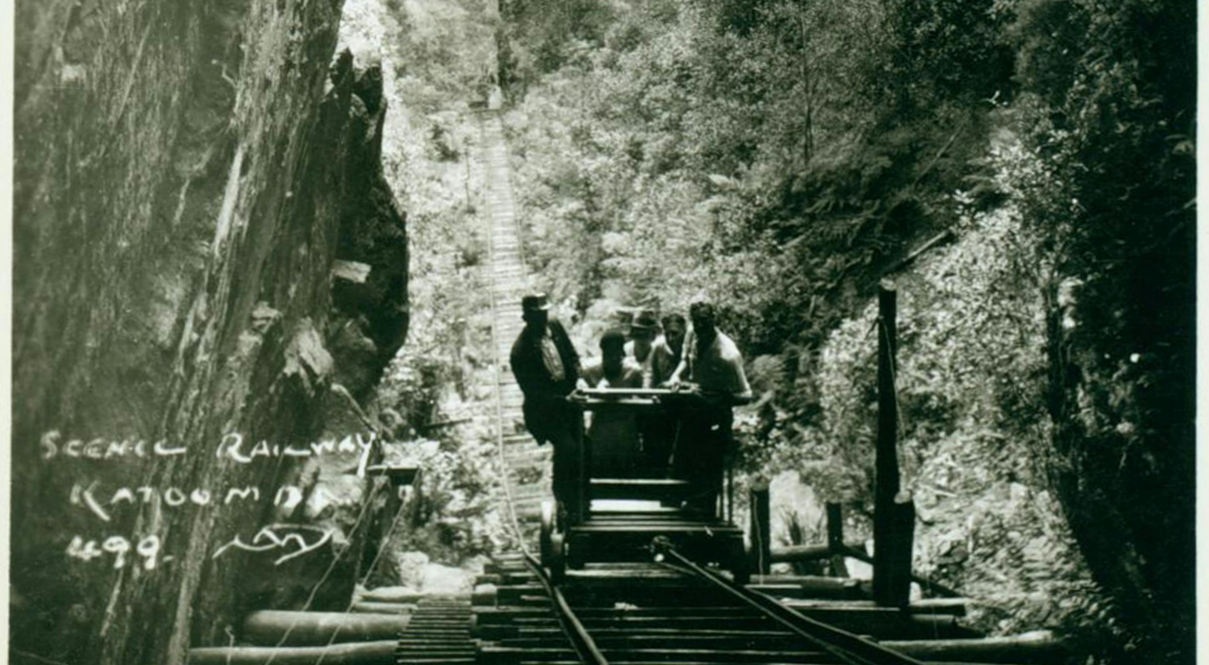 A vintage photograph shows a small group of people riding a scenic railway car through a narrow gorge surrounded by dense forest. The handwritten text Scenic Railway Katoomba 498 is visible in the lower left corner.