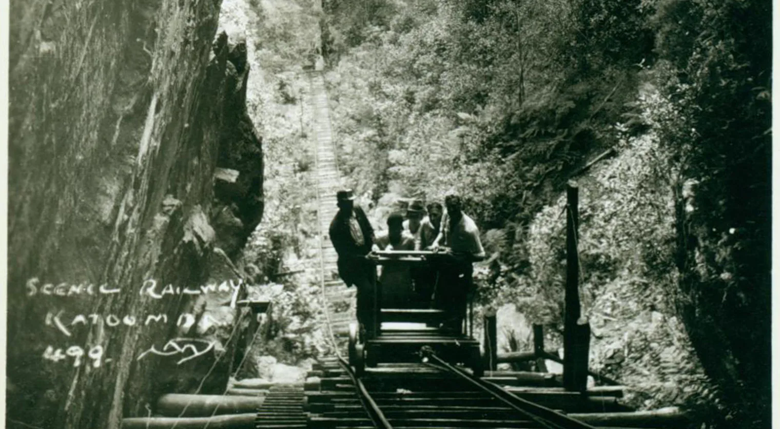 A vintage photograph shows a small group of people riding a scenic railway car through a narrow gorge surrounded by dense forest. The handwritten text Scenic Railway Katoomba 498 is visible in the lower left corner.
