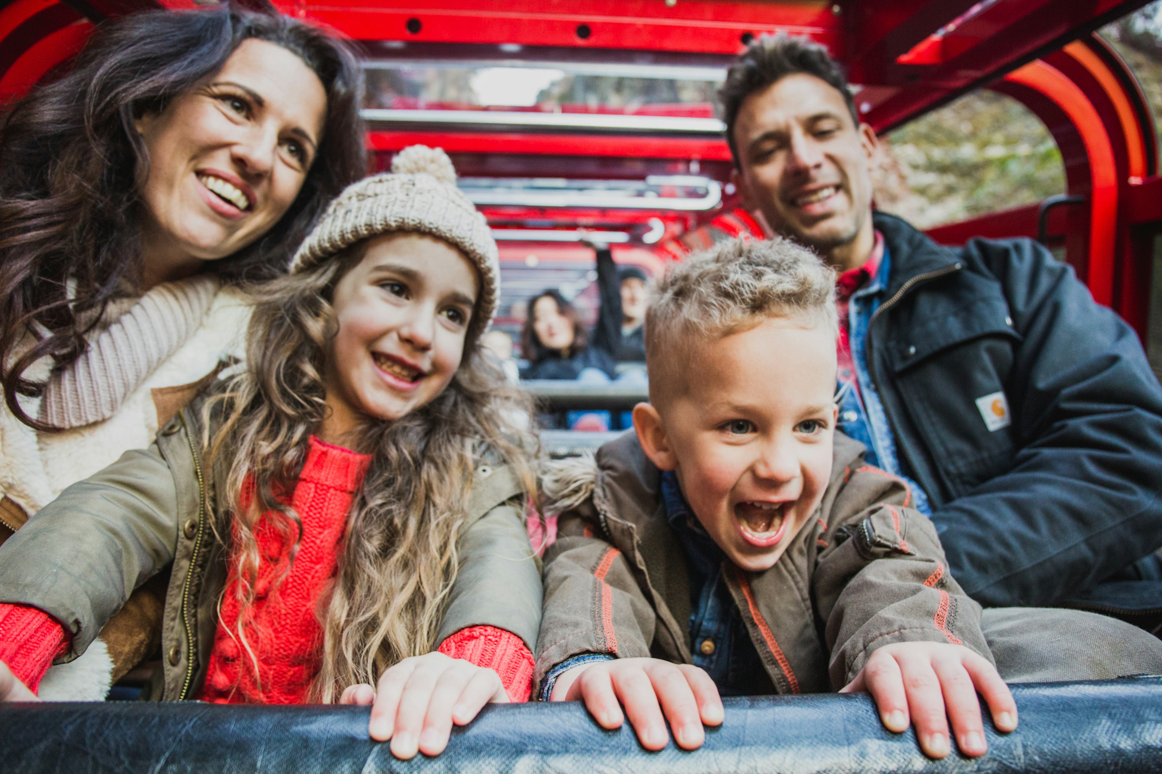 A family of four, including two children, is smiling and enjoying a ride in an open-top vehicle. They are dressed warmly in jackets and hats, appearing joyful and engaged with their surroundings. The vehicle is bright red.