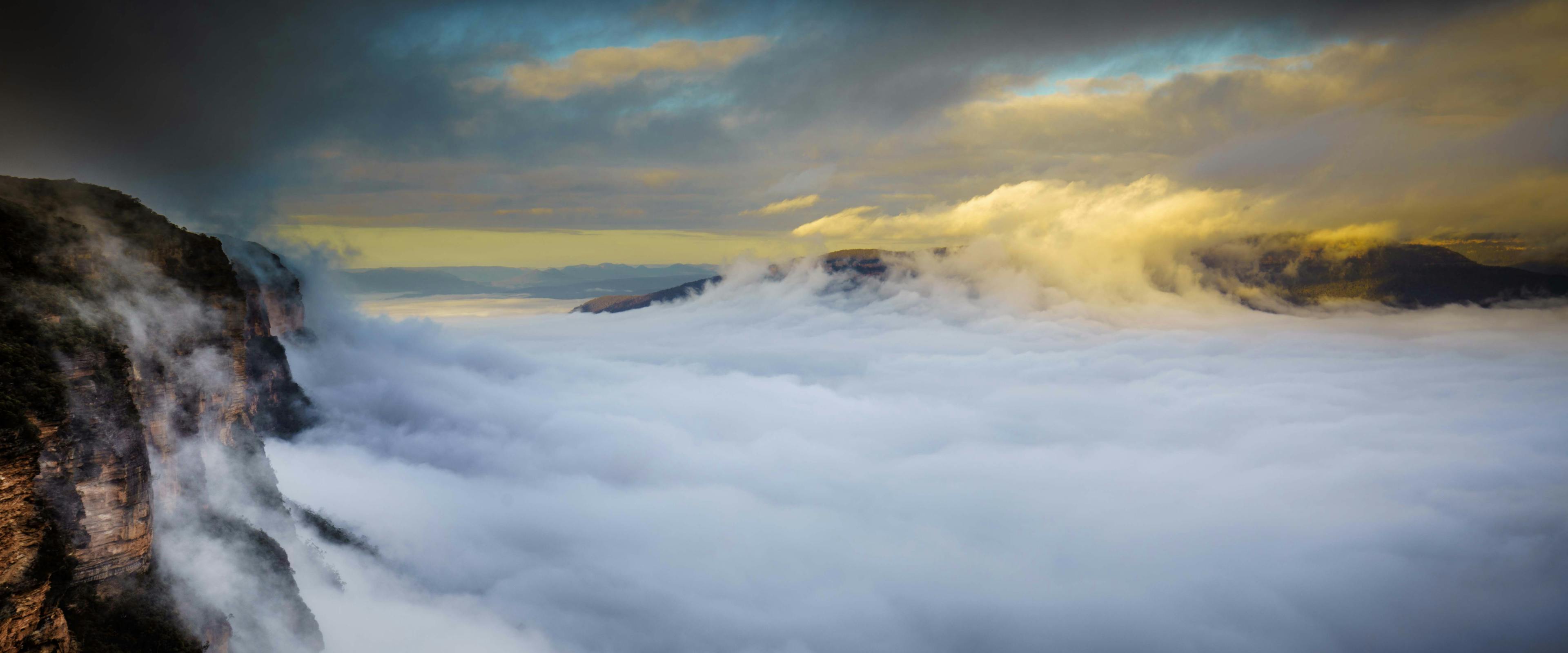 A dramatic landscape with thick clouds and mist covering a mountain range. The sky is partly cloudy with sunlight breaking through, casting a warm glow. A sheer cliffside is visible on the left, adding depth to the scene.