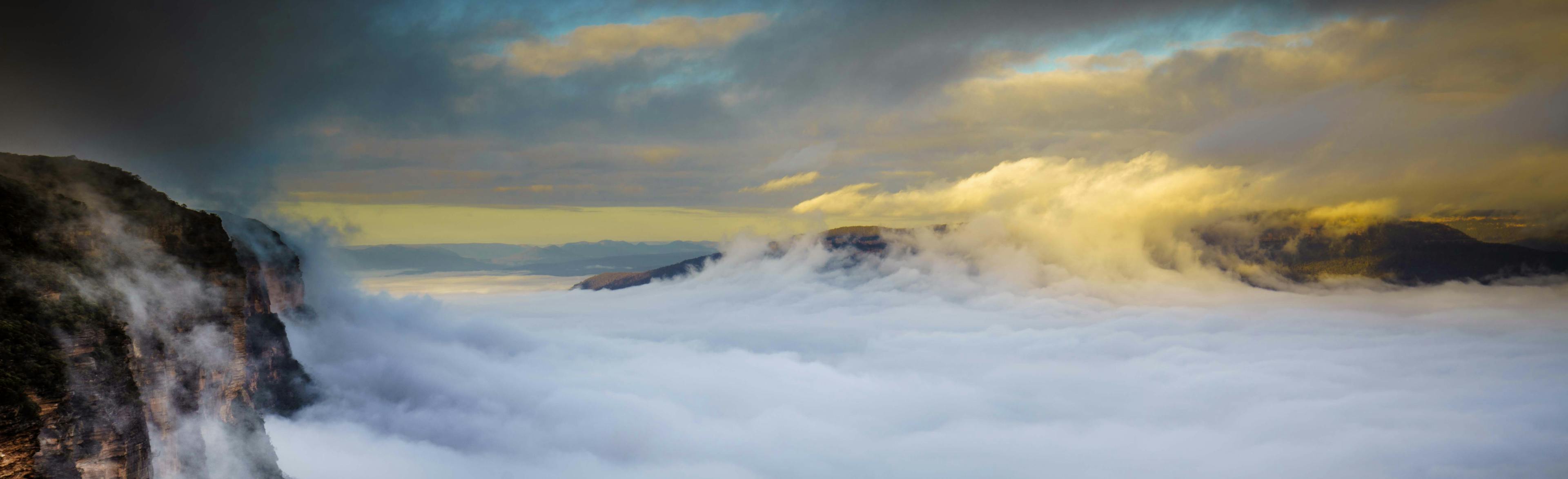 A dramatic landscape with thick clouds and mist covering a mountain range. The sky is partly cloudy with sunlight breaking through, casting a warm glow. A sheer cliffside is visible on the left, adding depth to the scene.