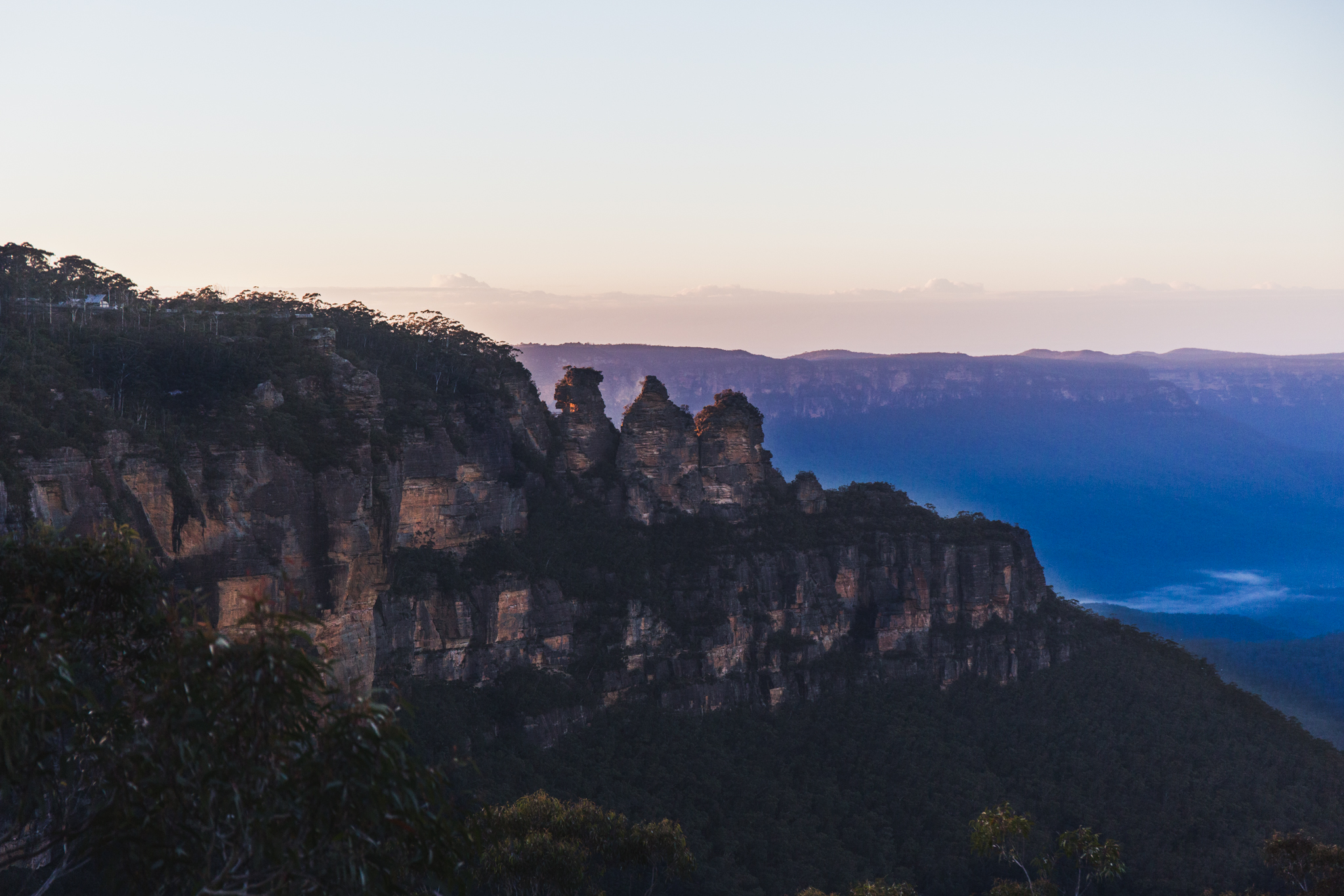 Scenic view of the Three Sisters rock formation at sunset in the Blue Mountains, Australia. The jagged peaks stand against a backdrop of lush forest and distant misty mountains, under a softly colored sky.