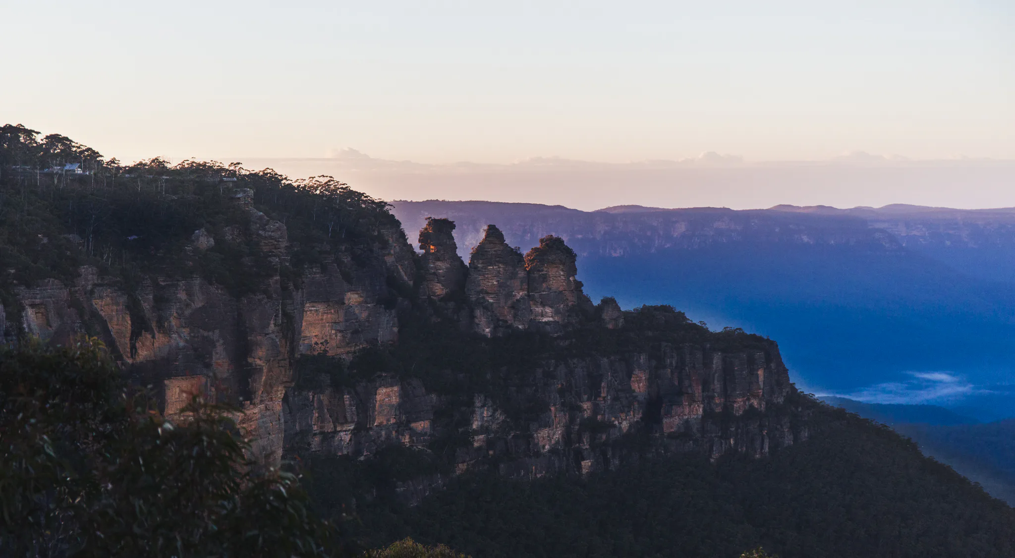 Scenic view of the Three Sisters rock formation at sunset in the Blue Mountains, Australia. The jagged peaks stand against a backdrop of lush forest and distant misty mountains, under a softly colored sky.