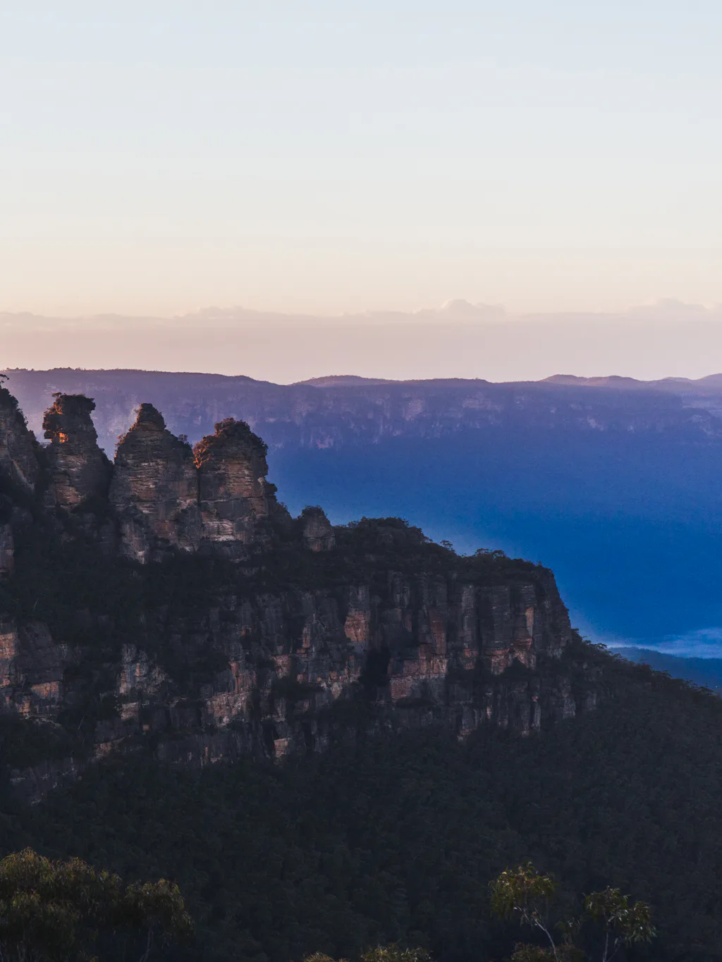 Scenic view of the Three Sisters rock formation at sunset in the Blue Mountains, Australia. The jagged peaks stand against a backdrop of lush forest and distant misty mountains, under a softly colored sky.