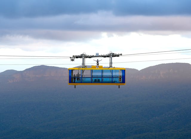 A person stands on top of a cable car suspended in mid-air with a mountainous landscape in the background. The sky is overcast, with clouds casting shadows on the distant hills and valleys.