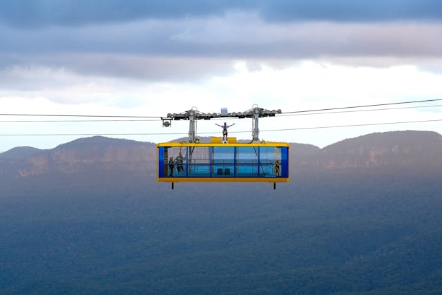 A person stands on top of a cable car suspended in mid-air with a mountainous landscape in the background. The sky is overcast, with clouds casting shadows on the distant hills and valleys.