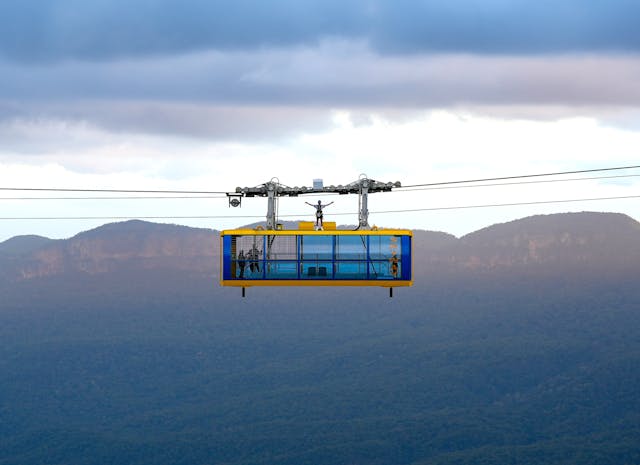 A person stands on top of a cable car suspended in mid-air with a mountainous landscape in the background. The sky is overcast, with clouds casting shadows on the distant hills and valleys.