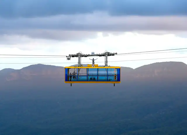 A person stands on top of a cable car suspended in mid-air with a mountainous landscape in the background. The sky is overcast, with clouds casting shadows on the distant hills and valleys.