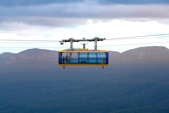 A person stands on top of a cable car suspended in mid-air with a mountainous landscape in the background. The sky is overcast, with clouds casting shadows on the distant hills and valleys.