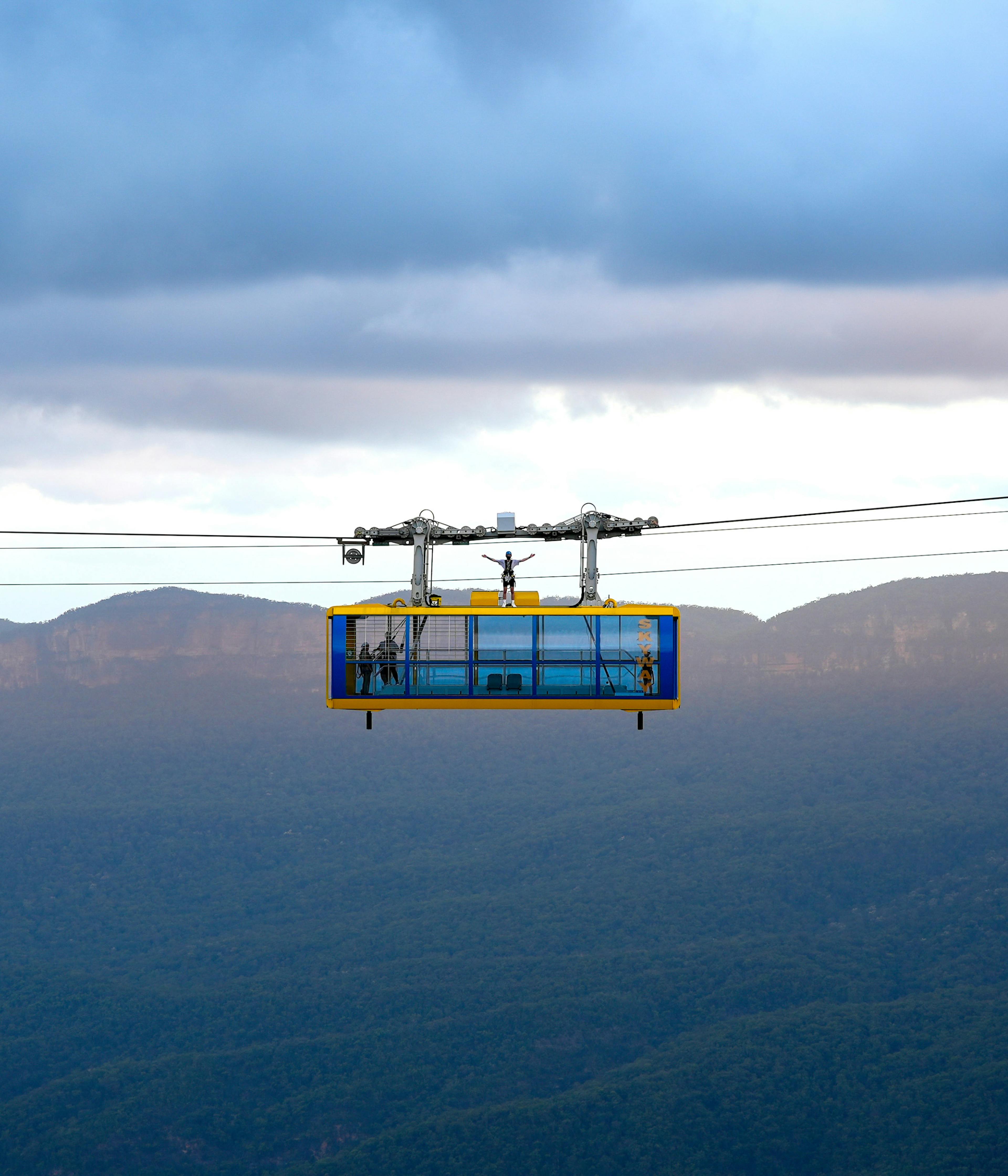 A person stands on top of a cable car suspended in mid-air with a mountainous landscape in the background. The sky is overcast, with clouds casting shadows on the distant hills and valleys.