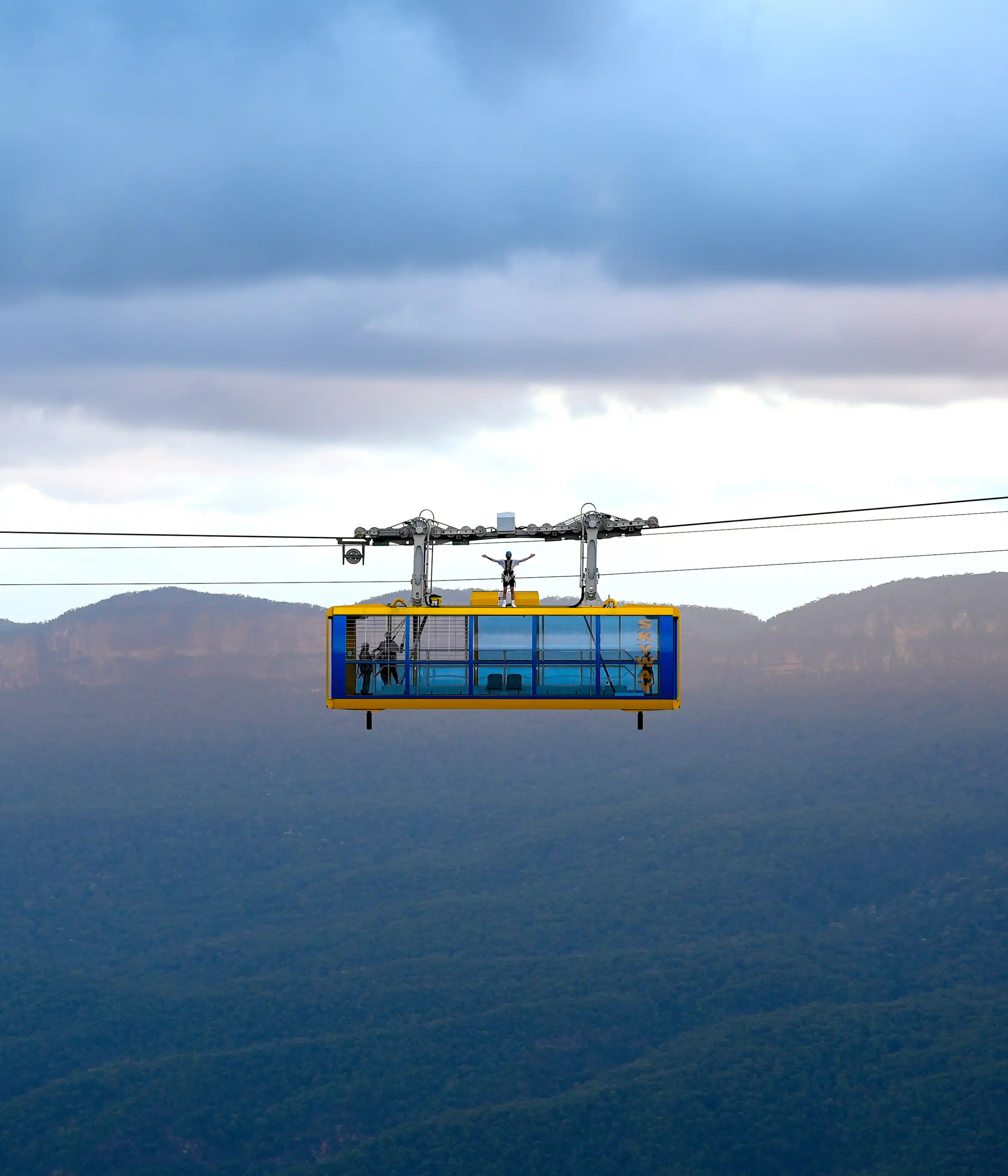 A person stands on top of a cable car suspended in mid-air with a mountainous landscape in the background. The sky is overcast, with clouds casting shadows on the distant hills and valleys.