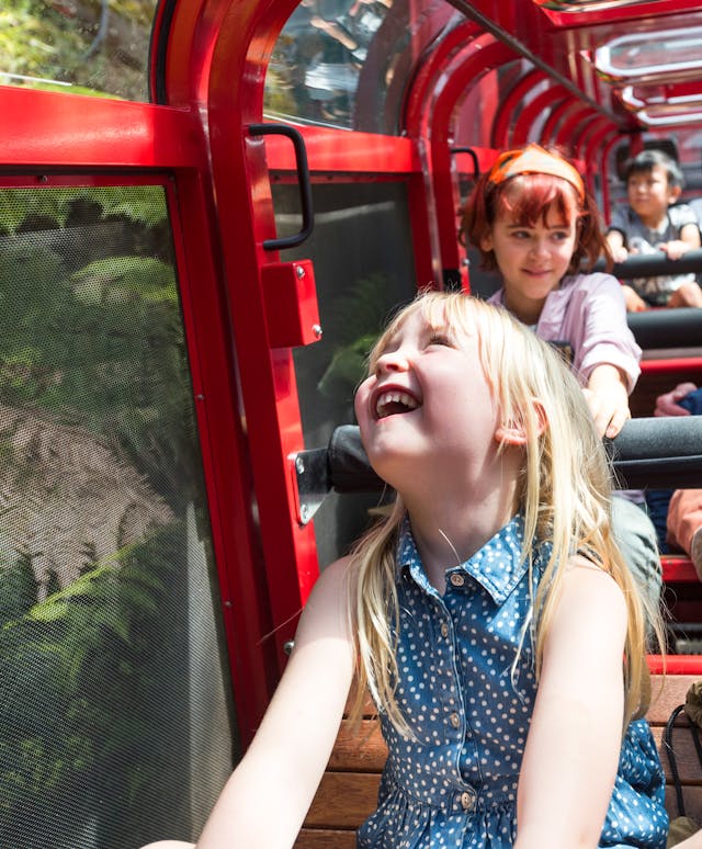 A group of people, mostly children, enjoying a ride in the railway. A young girl with blonde hair and a blue dress is laughing joyfully. Passengers are surrounded by lush green foliage and wearing casual, summer clothing.