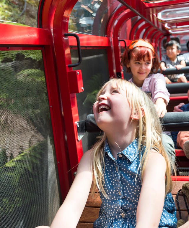 A group of people, mostly children, enjoying a ride in the railway. A young girl with blonde hair and a blue dress is laughing joyfully. Passengers are surrounded by lush green foliage and wearing casual, summer clothing.