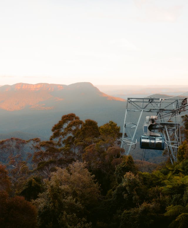 A cable car travels across a lush landscape with dense greenery and tall trees in the foreground. In the distance, vast rolling hills and a mountain range are illuminated by the golden light of sunset.