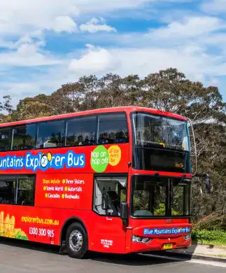 A red double-decker bus labeled Blue Mountains Explorer Bus is on a road with lush trees and mountains in the background. The scene is under a clear blue sky.