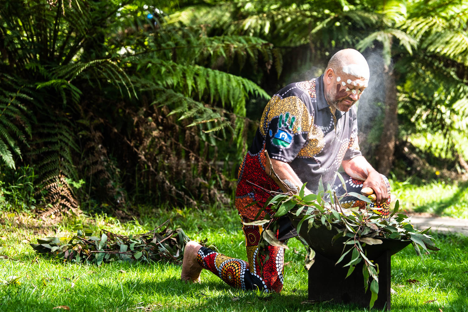 A person wearing colorful, patterned clothing performs a traditional ceremony outdoors, kneeling beside a smoking vessel filled with leaves. Tall green ferns and grass surround the scene.