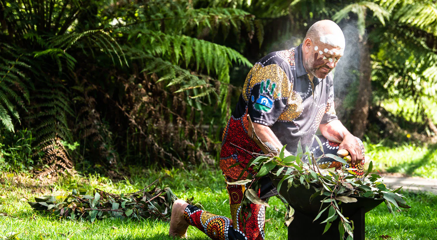 A person wearing colorful, patterned clothing performs a traditional ceremony outdoors, kneeling beside a smoking vessel filled with leaves. Tall green ferns and grass surround the scene.