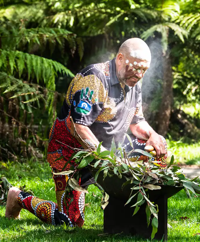 A person wearing colorful, patterned clothing performs a traditional ceremony outdoors, kneeling beside a smoking vessel filled with leaves. Tall green ferns and grass surround the scene.