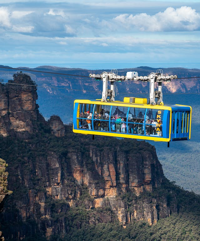 A cable car with tourists travels across a lush, mountainous landscape with rocky cliffs in the background, under a cloudy sky. The area is covered in dense green foliage.