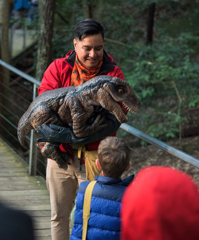 A man in a red jacket holds a baby dinosaurt, entertaining a child in a blue jacket on a wooden walkway. Other people in colourful jackets stand nearby. The background is lush with green trees.