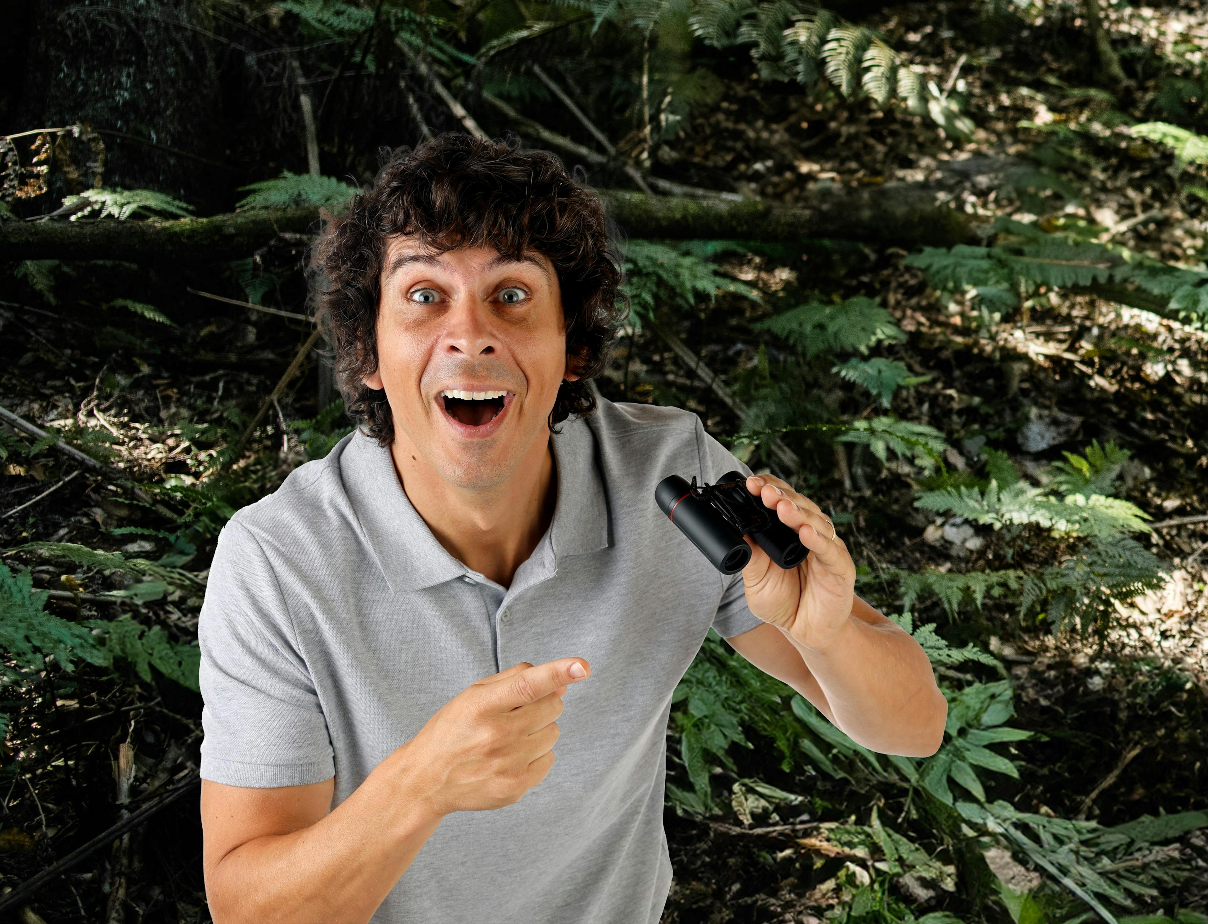 A person with curly hair and a surprised expression points at a pair of binoculars in their hand. They are wearing a grey polo shirt and standing amidst lush greenery, with branches and leaves in the background. The person is Andy Day. 