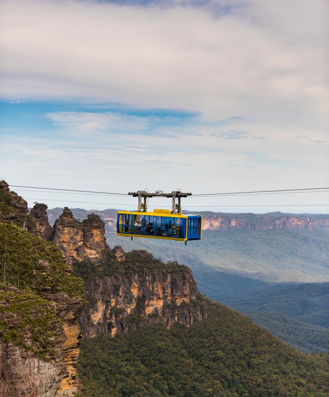 A cable car travels across a valley with lush green forests and the Three Sisters. The sky is partly cloudy, and the landscape stretches into the distance, showcasing a dramatic and picturesque view.