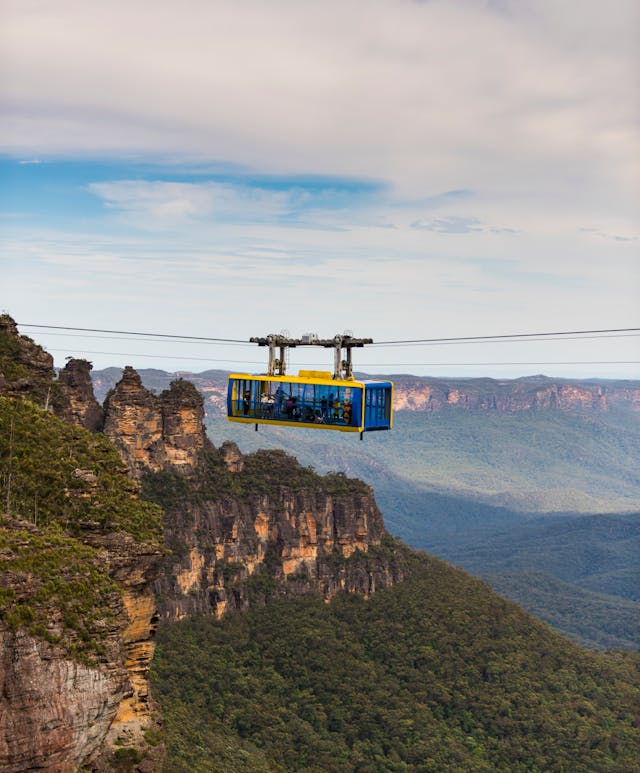 A cable car travels across a valley with lush green forests and the Three Sisters. The sky is partly cloudy, and the landscape stretches into the distance, showcasing a dramatic and picturesque view.