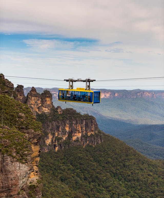 A cable car travels across a valley with lush green forests and the Three Sisters. The sky is partly cloudy, and the landscape stretches into the distance, showcasing a dramatic and picturesque view.