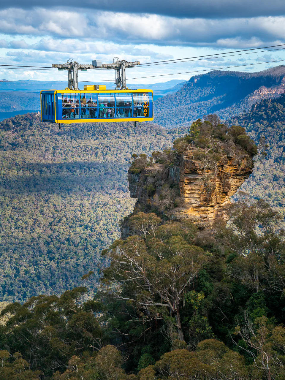 A cable car glides above lush forests and rugged cliffs, with a prominent rock formation below. The expansive landscape features a range of greenery and distant mountains under a cloudy sky.