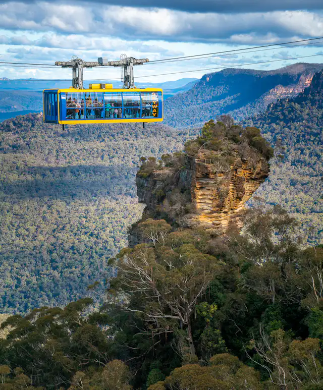 A cable car glides above lush forests and rugged cliffs, with a prominent rock formation below. The expansive landscape features a range of greenery and distant mountains under a cloudy sky.
