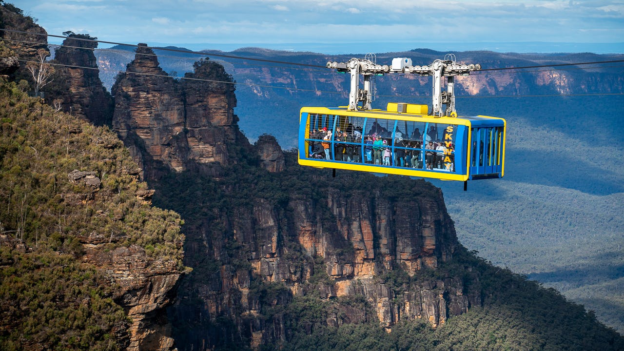 A cable car with tourists travels across a lush, mountainous landscape with rocky cliffs in the background, under a cloudy sky. The area is covered in dense green foliage.