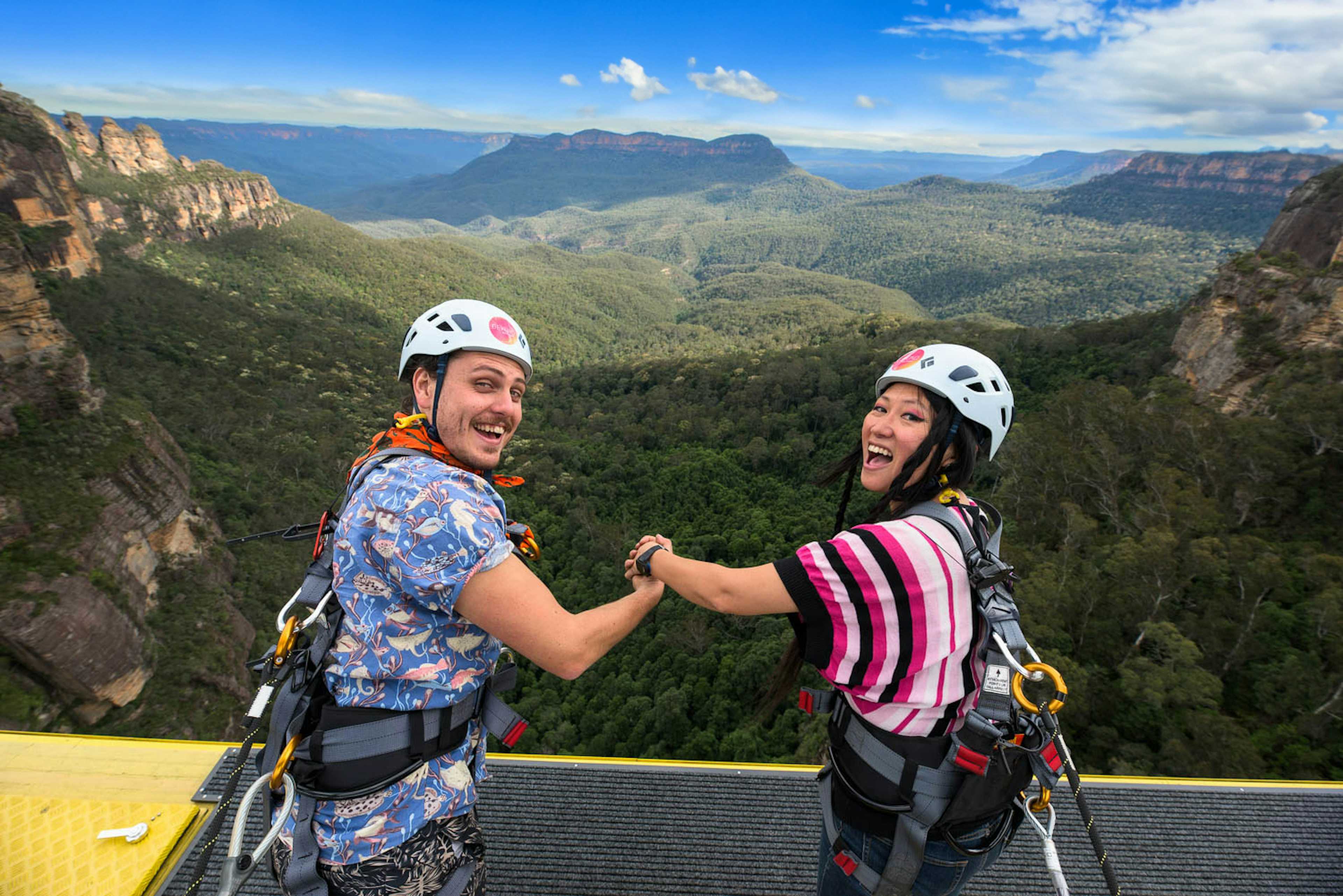 A man and woman wearing helmets and harnesses hold hands and smile at the edge of the Skyway cable car with a vast forested valley and mountain range in the background. The sky is clear with a few clouds.