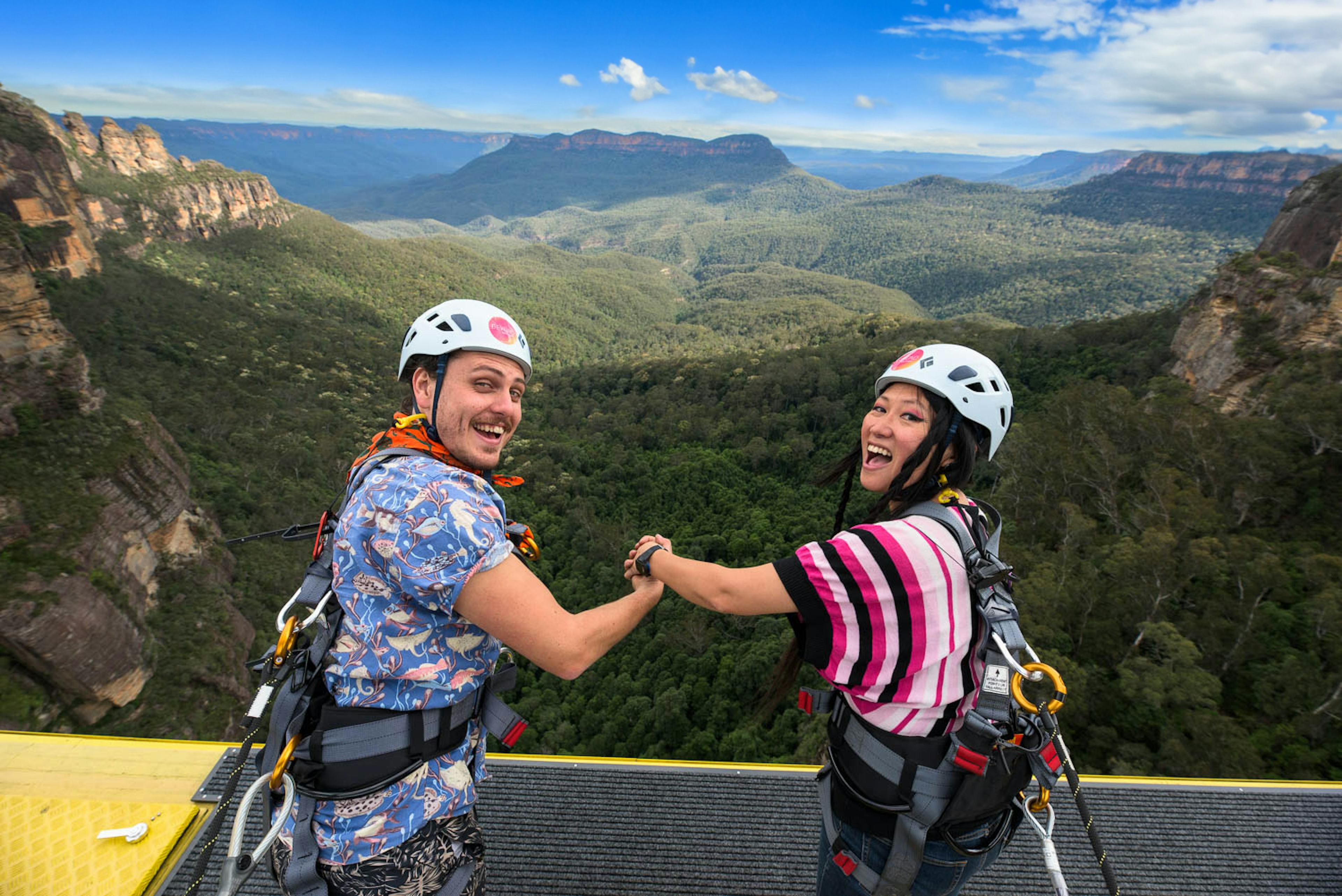 A man and woman wearing helmets and harnesses hold hands and smile at the edge of the Skyway cable car with a vast forested valley and mountain range in the background. The sky is clear with a few clouds.